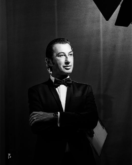 Uomo in abito elegante con papillon, braccia incrociate, sfondo scuro.