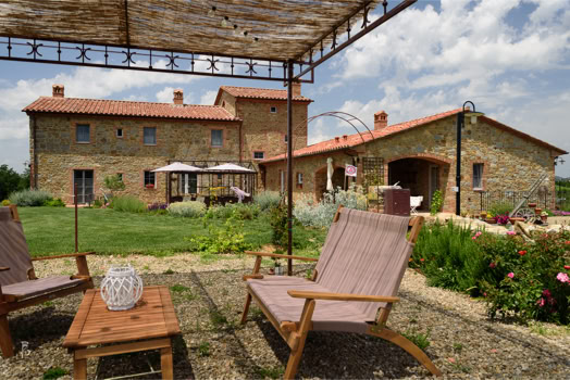 Giardino con sedie a sdraio e tavolino sotto una pergola, sullo sfondo una casa in pietra con tetto in tegole rosse.