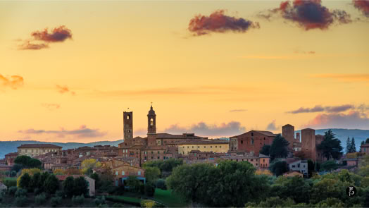 Veduta di una città con edifici storici e torri al tramonto, circondata da alberi e colline, con un cielo colorato da nuvole sparse.