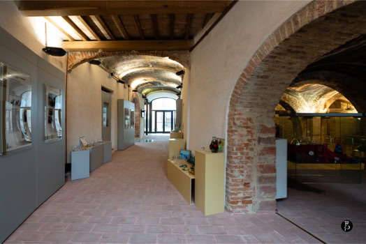 Corridoio con soffitto a travi di legno, pareti in mattoni e archi, con esposizioni su entrambi i lati.