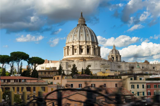 Cupola di una grande basilica con edifici storici circostanti e alberi, sotto un cielo parzialmente nuvoloso.