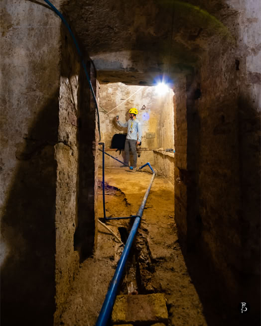 Una persona con casco giallo cammina in un tunnel sotterraneo con pareti di pietra e un tubo blu sul pavimento.