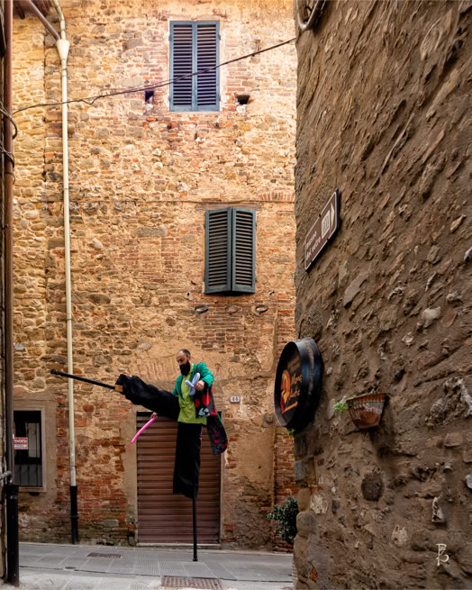 Edificio in mattoni con finestre chiuse, una figura sospesa su un bastone, e una strada stretta.