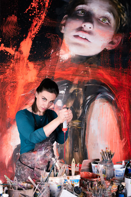 Artista che lavora in uno studio con un grande dipinto di un volto sullo sfondo, circondata da pennelli e materiali artistici.