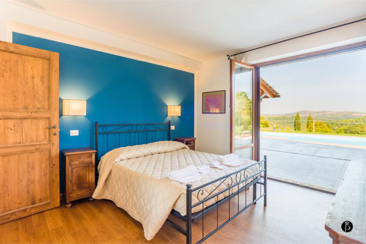 Camera da letto con parete blu, letto matrimoniale con copriletto chiaro, due comodini in legno, porta in legno a sinistra e grande finestra aperta con vista su un paesaggio esterno.