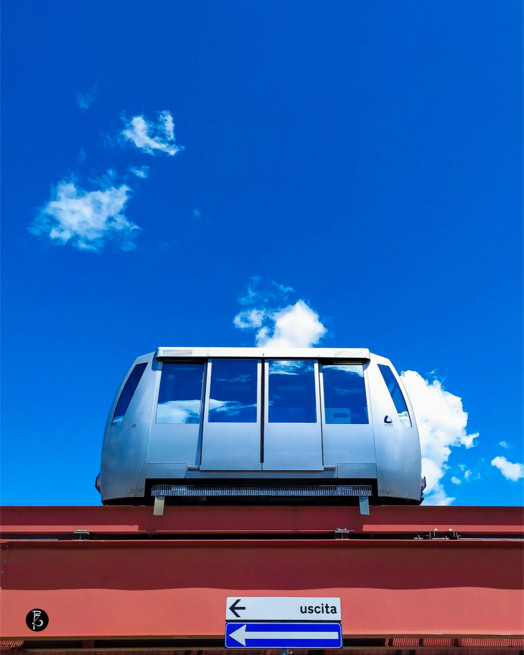 Un vagone di metropolitana sopraelevata su un binario rosso, con cielo blu e nuvole sullo sfondo.