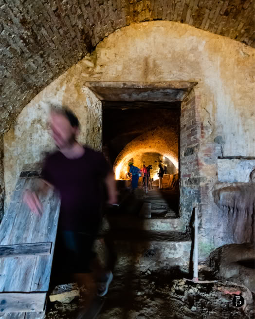 Persona sfocata che cammina in un corridoio di pietra con archi, illuminato da una luce calda in fondo.