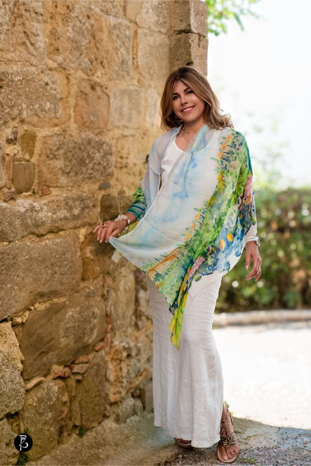 Donna in piedi accanto a un muro di pietra, indossa abiti bianchi e un foulard colorato.
