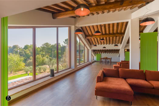 Interno di una stanza con ampie finestre, un soffitto con travi in legno, un divano arancione e un tavolo sullo sfondo.