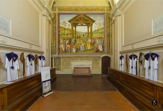Interno di una cappella con un grande affresco religioso sulla parete di fondo, panche di legno lungo i lati e abiti bianchi appesi.