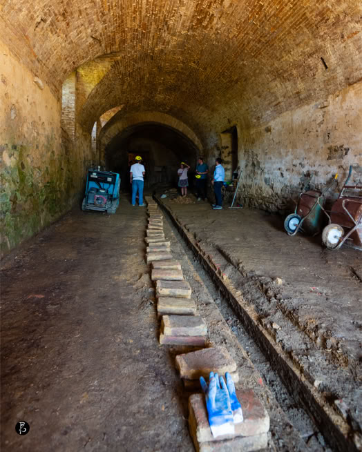 Interno di un tunnel con binari al centro, persone con caschi da lavoro, attrezzature e carrelli lungo i lati.