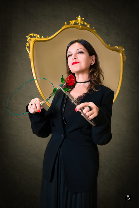 Donna in abito nero con una rosa rossa in mano, davanti a una cornice dorata.