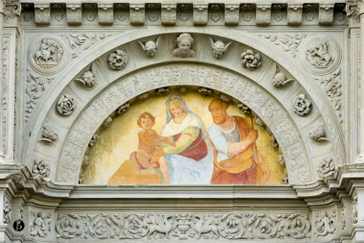 Affresco raffigurante tre figure umane, due adulti e un bambino, incorniciato da un arco decorato con motivi floreali e dettagli scolpiti.