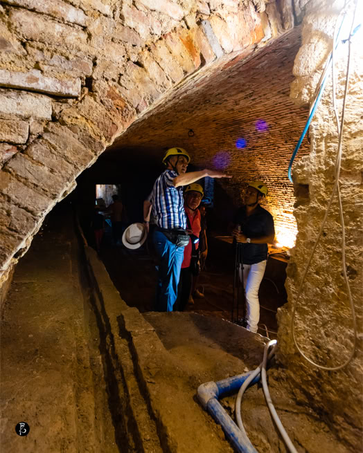 Persone in un tunnel di mattoni con caschi protettivi, una persona indica qualcosa.