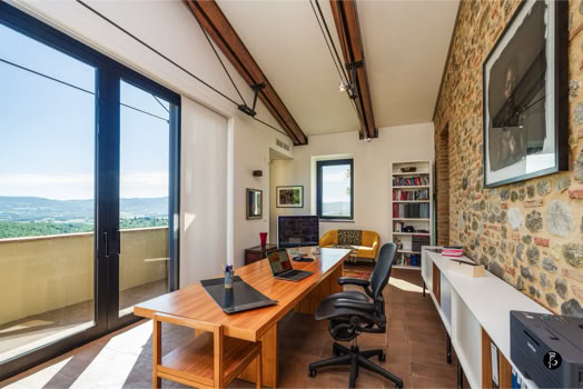 Ufficio moderno con scrivania in legno, sedie da ufficio, computer e scaffali. Parete in pietra a destra, grande finestra a sinistra con vista panoramica.