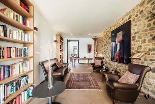 Sala con parete in pietra, libreria a sinistra, due poltrone in pelle marrone, tavolino rotondo, tappeto rosso e scrivania in fondo.