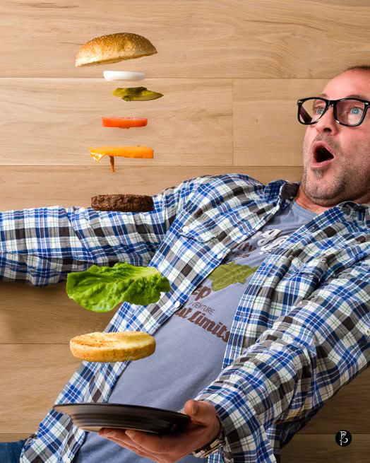Uomo con camicia a quadri e occhiali sorpreso mentre tiene un piatto, con ingredienti di hamburger sospesi in aria.