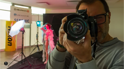 Uomo che scatta una foto con una fotocamera, sullo sfondo una persona in posa con luci colorate e attrezzatura fotografica.