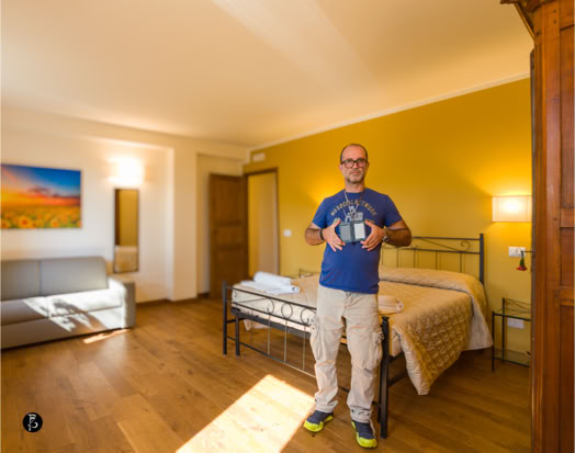 Uomo in piedi in una stanza con pavimento in legno, letto singolo, divano grigio, parete gialla e quadro colorato.