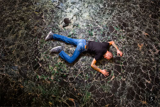 Una persona distesa a terra su un pavimento di pietre, indossa jeans e una maglietta scura.