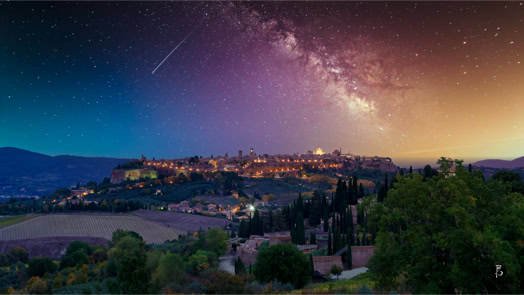 Veduta notturna di una città collinare illuminata, con la Via Lattea visibile nel cielo stellato sopra.