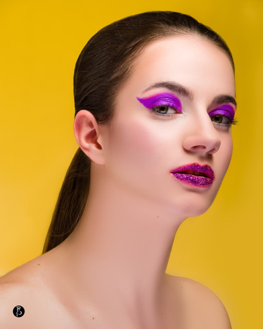 Donna con trucco occhi e labbra viola su sfondo giallo.