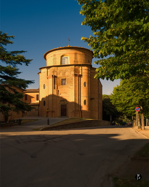 Edificio storico in mattoni con forma cilindrica, circondato da alberi verdi, sotto un cielo blu.