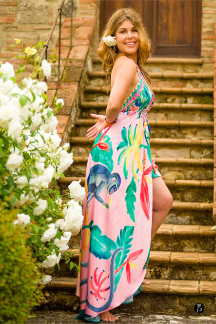 Donna in abito colorato con motivi floreali, in piedi su una scalinata di pietra, con fiori bianchi a sinistra.