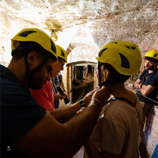 Persone con caschi gialli in una grotta, una persona sta aiutando un'altra con l'attrezzatura.