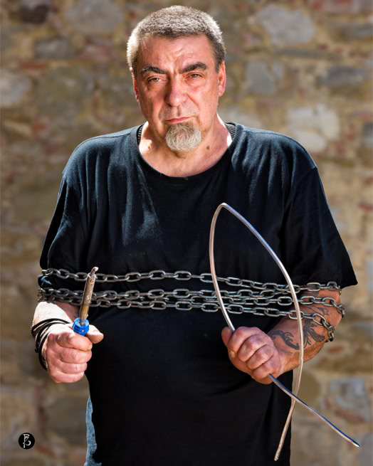 Uomo con barba e baffi, indossa una maglietta nera, avvolto in catene di metallo, tiene un cacciavite e un filo di metallo. Sfondo di muro di pietra.