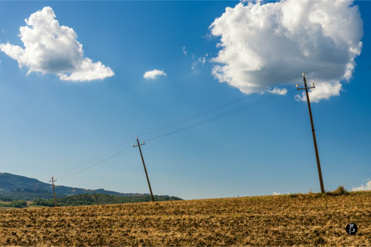Campo arato con due pali della luce inclinati, cielo azzurro con nuvole bianche.