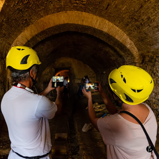 Due persone con caschi gialli scattano foto con i loro telefoni in un ambiente sotterraneo con archi in mattoni.