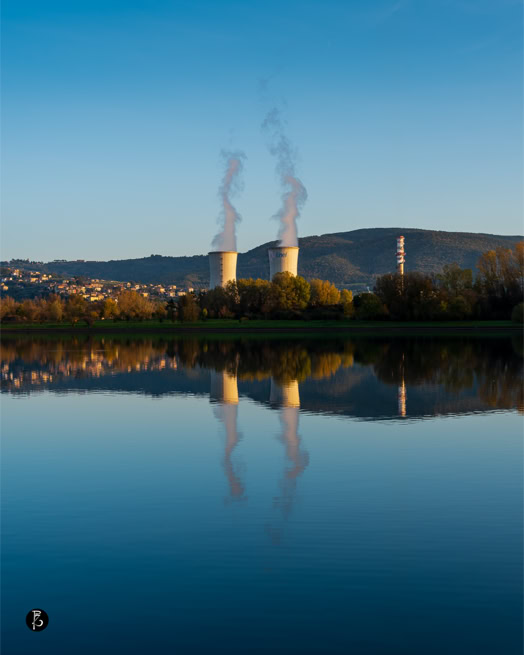 Due torri di raffreddamento che emettono vapore, con il loro riflesso visibile in un lago calmo, circondate da colline e vegetazione.