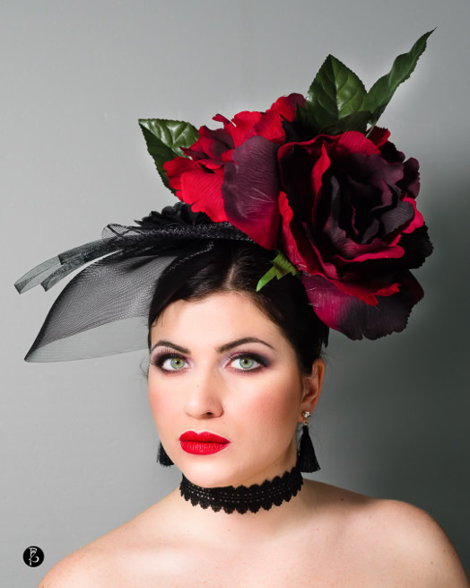 Donna con rossetto rosso e trucco marcato, indossa un cappello con grandi fiori rossi e foglie verdi.