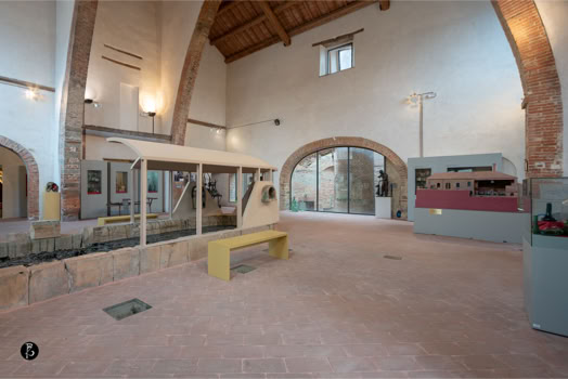 Interno di un edificio con soffitto alto e travi in legno, pareti bianche, pavimento in mattoni, un arco in mattoni, una panca gialla e vetrine espositive.
