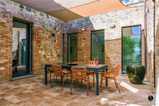 Cortile con tavolo e sedie in legno, pavimento in pietra, pareti in mattoni e pietra, vasi decorativi e tende da sole.