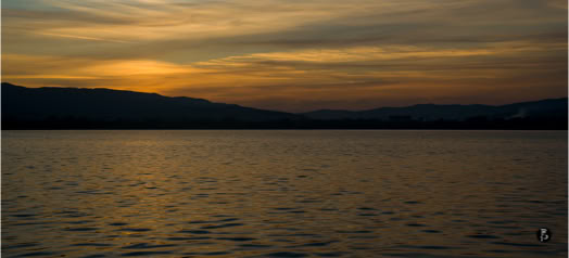 Tramonto su un lago con colline all'orizzonte e cielo nuvoloso.
