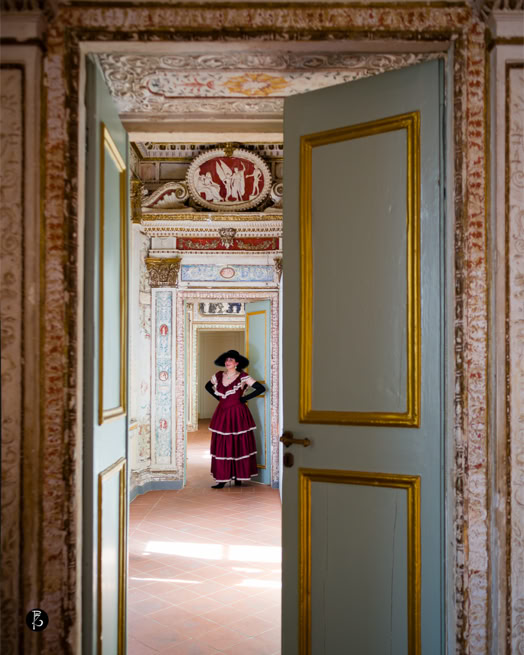 Porta aperta che mostra una stanza decorata con affreschi, una persona in abito rosso e cappello si trova in fondo alla stanza.
