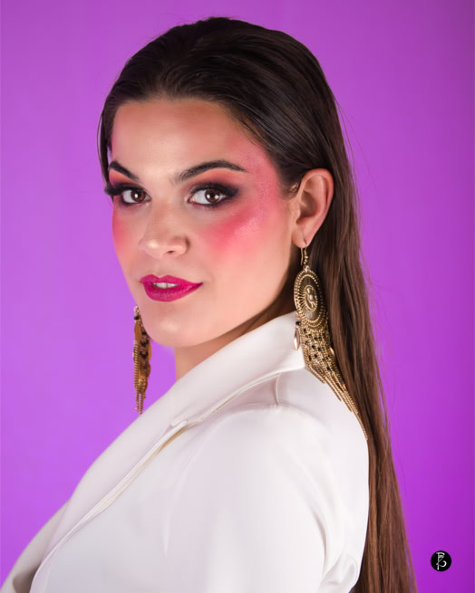 Donna con capelli lunghi e scuri, indossa un abito bianco e orecchini dorati, su sfondo viola.