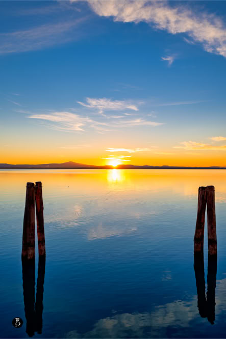 Tramonto su un lago calmo con due pali di legno in primo piano e cielo parzialmente nuvoloso.