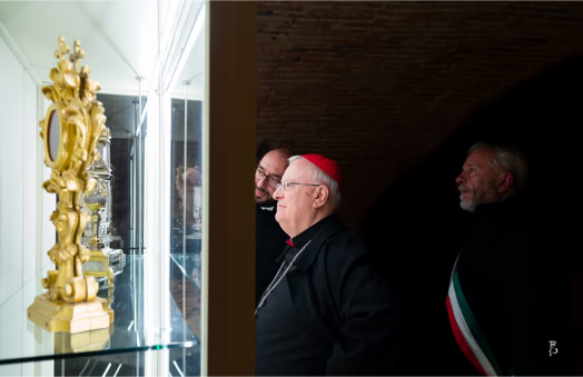 Tre uomini osservano una scultura dorata all'interno di una teca di vetro. Uno degli uomini indossa un copricapo rosso.