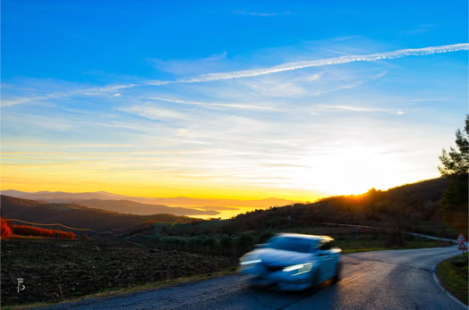 Auto in movimento su una strada di campagna al tramonto, con colline e cielo blu sullo sfondo.