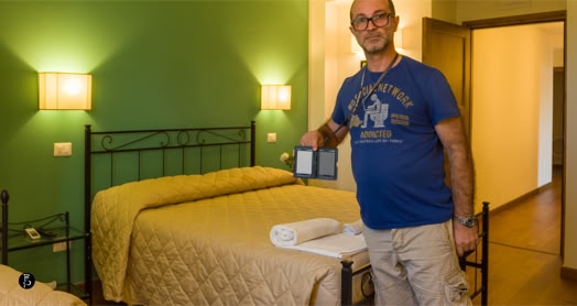 Uomo in piedi accanto a un letto in una stanza, con pareti verdi e due lampade accese.