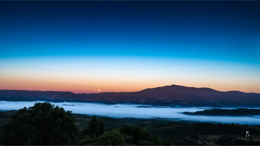 Paesaggio montano al tramonto con cielo sfumato dal blu all'arancione, una catena montuosa all'orizzonte e una valle coperta di nebbia.