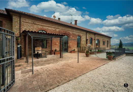 Edificio in mattoni con tetto in tegole, portico con sedie e tavolo, cielo nuvoloso sullo sfondo.