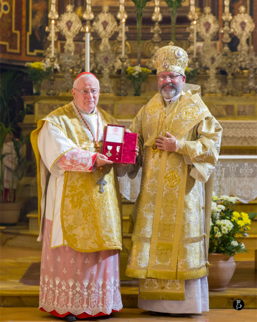 Due uomini in abiti religiosi dorati, uno dei quali tiene una scatola rossa, in piedi davanti a un altare ornato con candele e fiori.