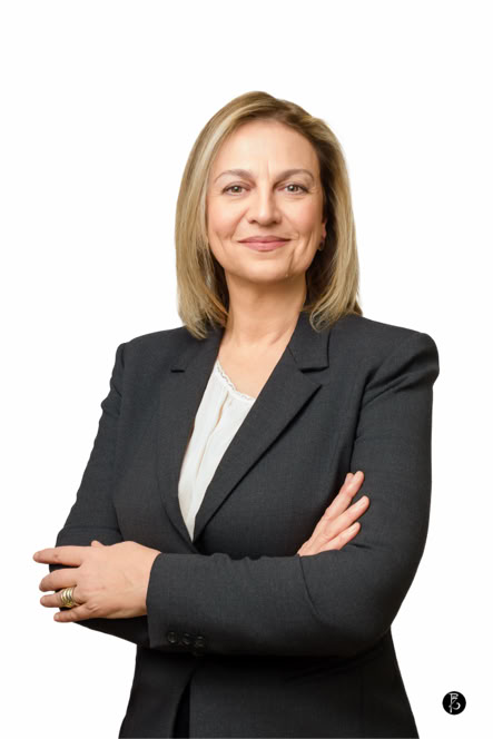 Donna con capelli biondi, in abito formale scuro, con le braccia incrociate, su sfondo bianco.