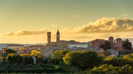 Panorama di una città con torri medievali al tramonto, circondata da alberi e colline.