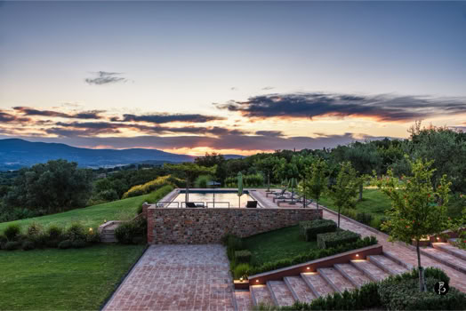 Vista di un giardino con piscina, circondato da alberi e vegetazione, con un cielo al tramonto e montagne sullo sfondo.