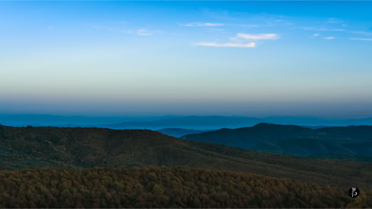 Paesaggio collinare al tramonto con cielo azzurro e alcune nuvole sottili.